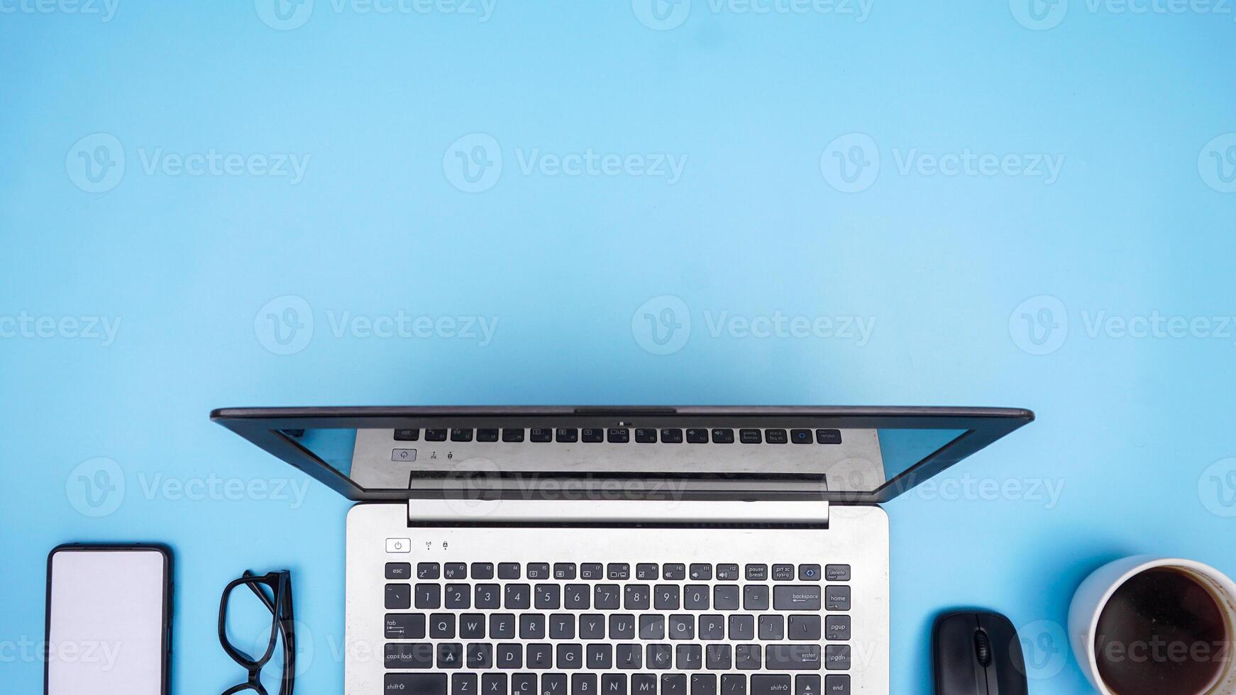 Top View of Working Space with Laptop, Mouse, Coffee Cup, Smartphone, and Glasses on Blue Background photo