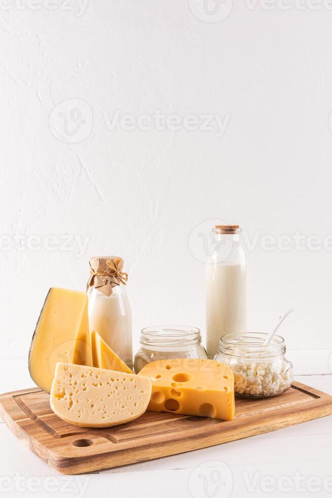 Many varieties of cheese on a board, bottles of milk, cottage cheese, sour cream on a light table. The holiday of Shavuot. Vertical view. photo