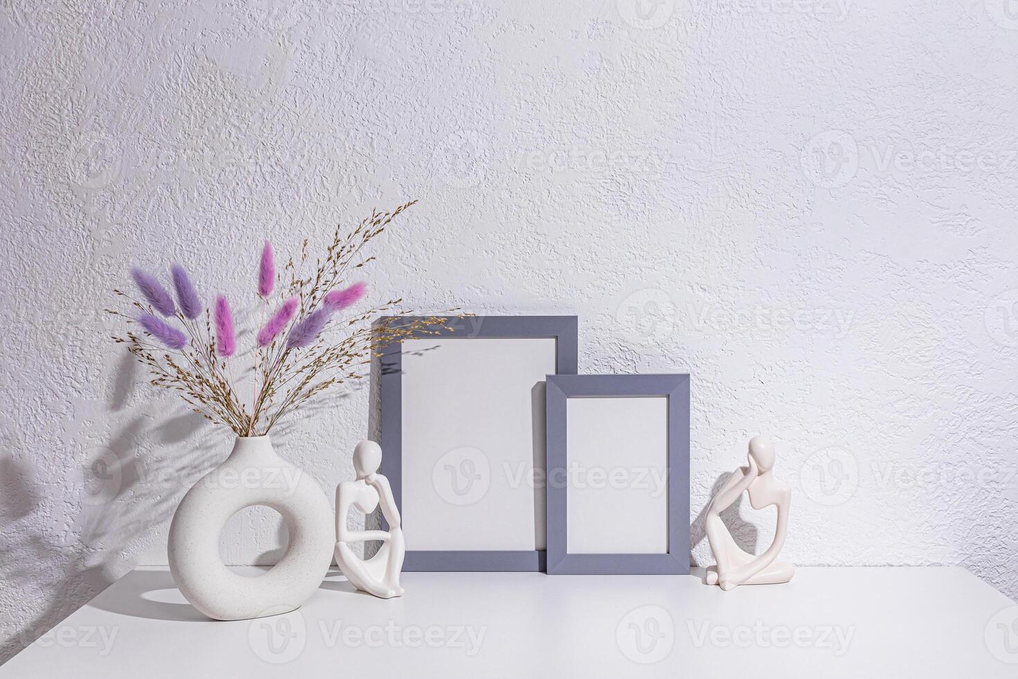 Two gray photo frames with empty space, a stylish vase with lilac dried flowers on a white table near the white wall. Beautiful interior of the room.