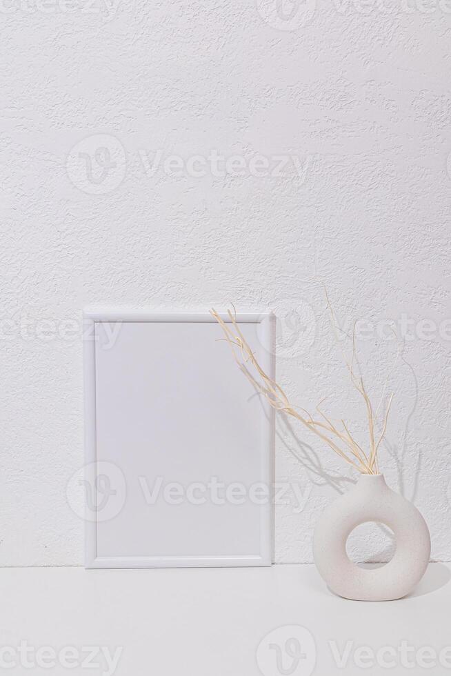 Blank photo frame mockup standing on the white table or chest. a stylish vase with dry branches. Front view. White textured wall. interior