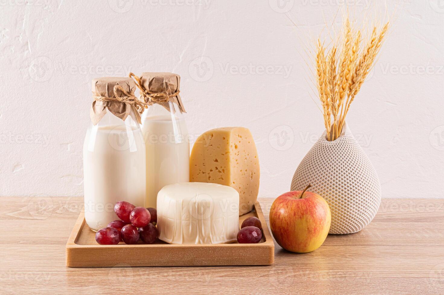 cheese, two bottles of milk, grapes, an apple, a vase with ears of wheat on a wooden table. Traditional treats and symbols of the Shavuot. Front view. photo