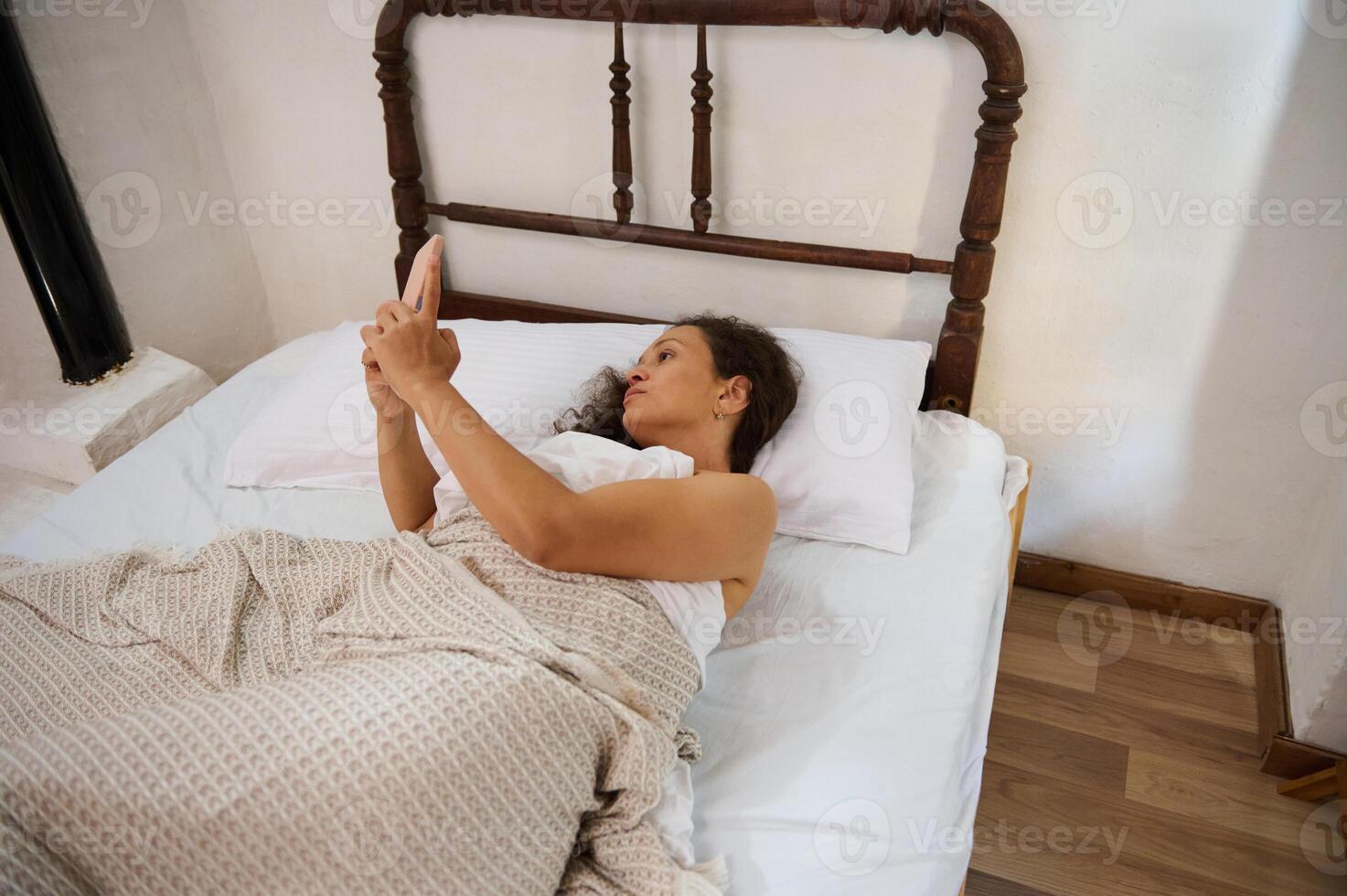 Woman relaxing in bed while browsing her smartphone photo