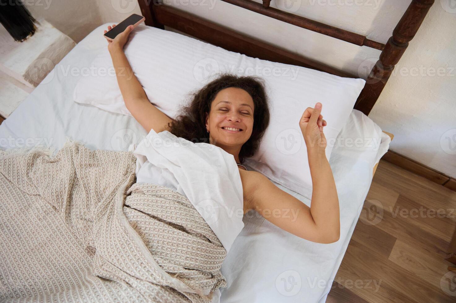 Woman waking up happily in bed holding a phone and smiling joyously photo
