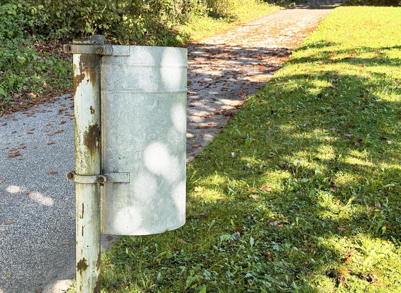 Rusty metal trash can on green park path with autumn leaves photo