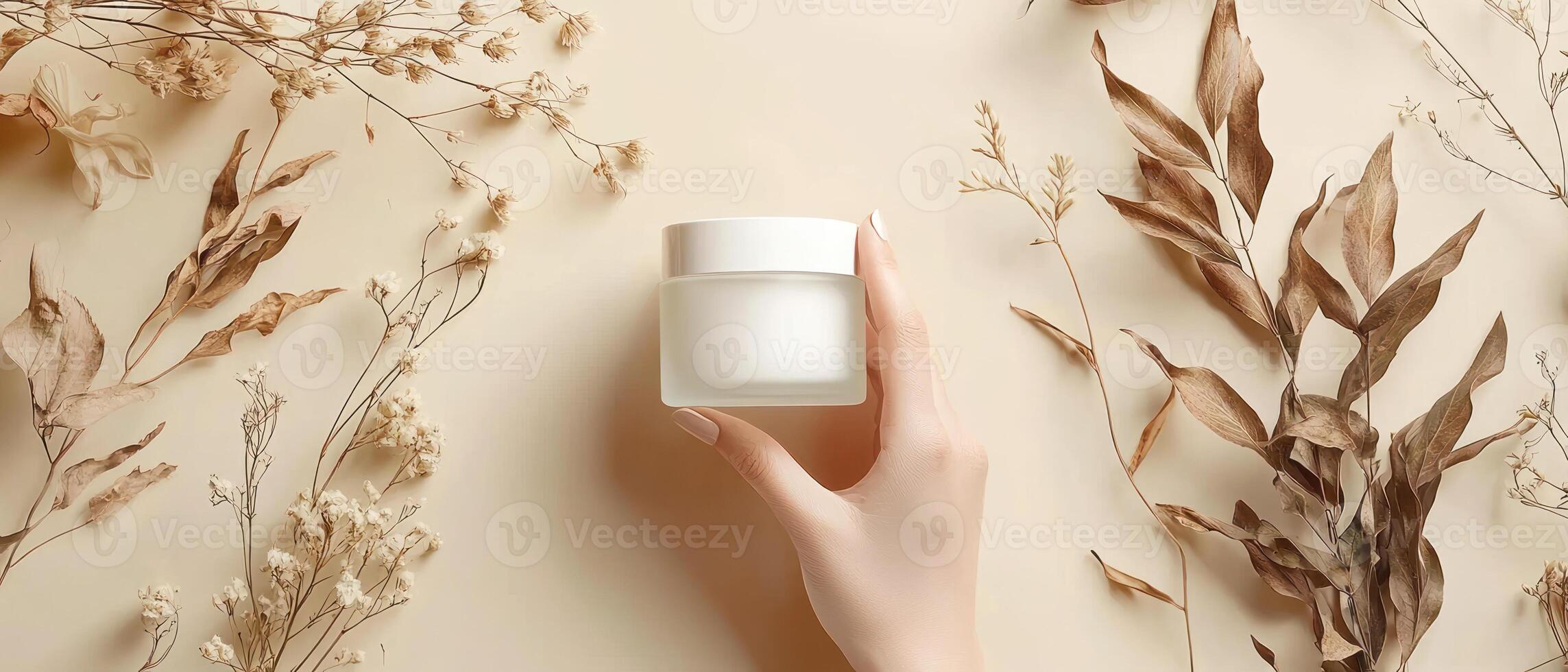 hand holding frosted glass cream jar surrounded by dried flowers and leaves on beige background creates serene atmosphere photo