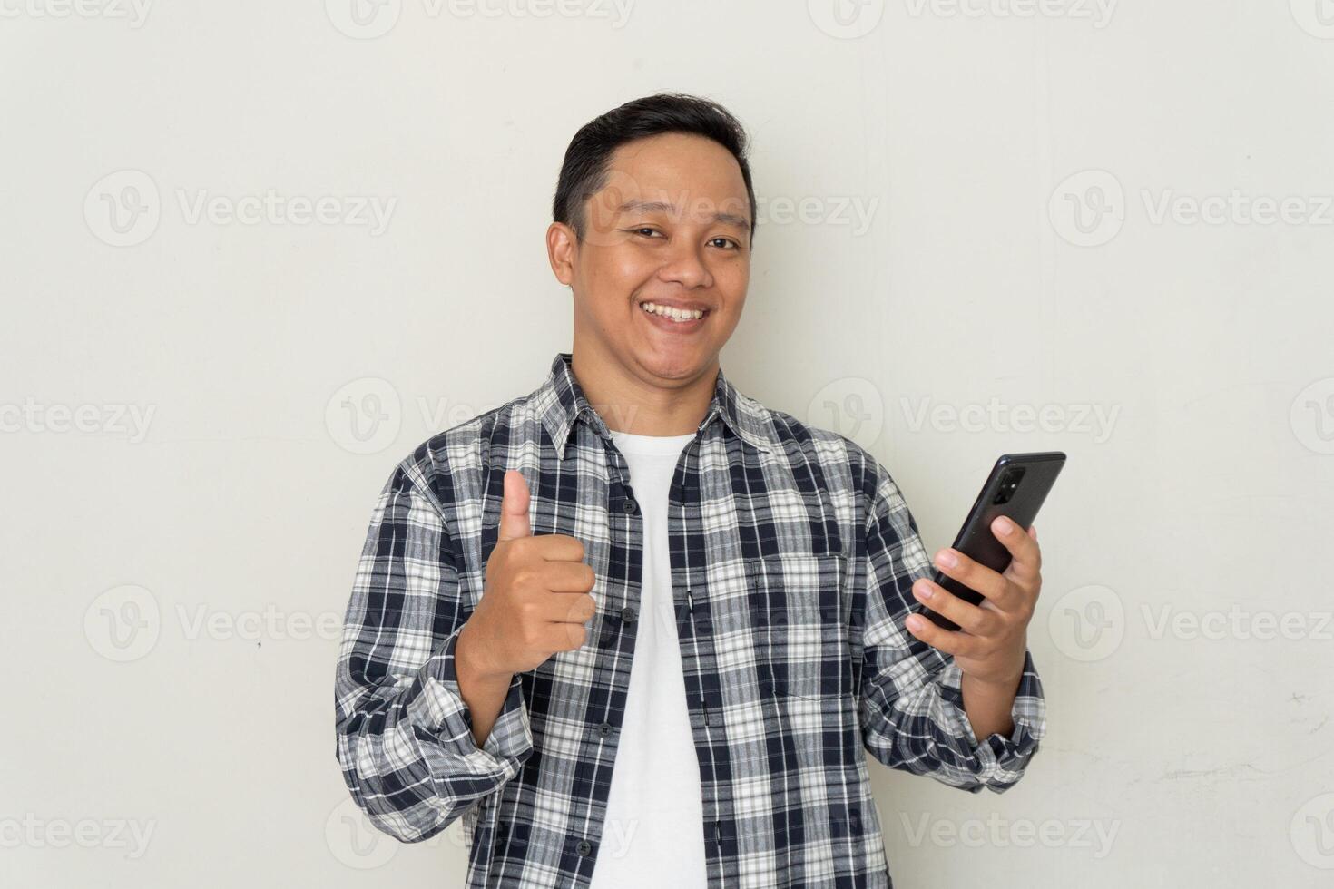 Young Asian man smiling happy and showing thumbs up gesture while holding his mobile phone. photo