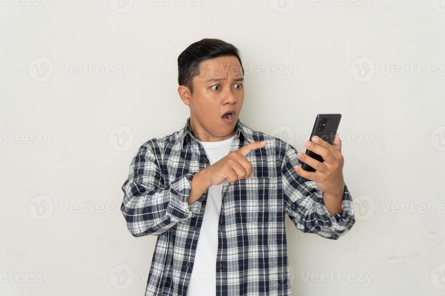 Young Asian man pointing to mobile phone with afraid and surprised expression. Promo and bad news concept photo
