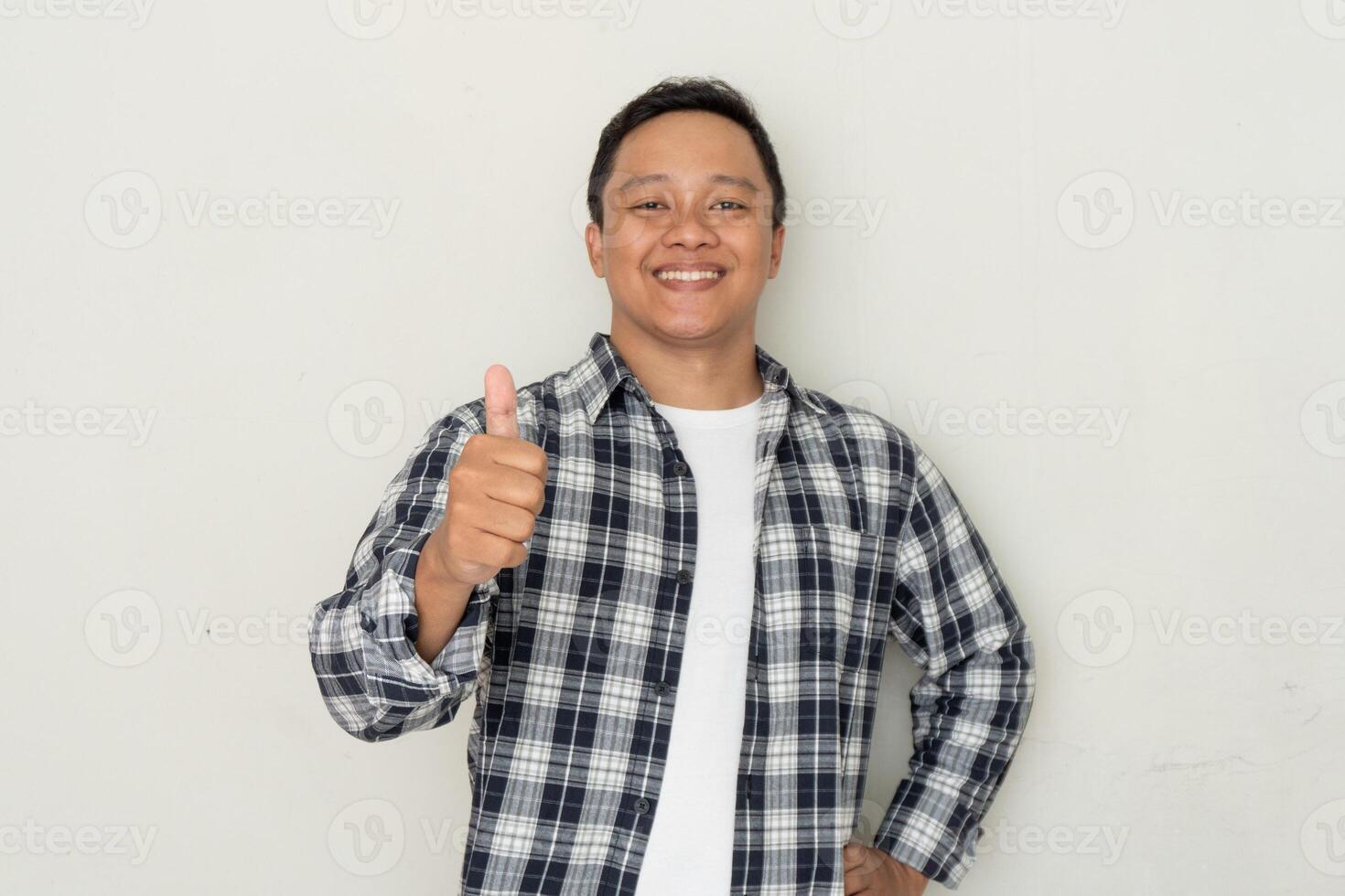 Attractive young Asian man gives a thumbs up and smiles confidently over white background. Concept of positivity, approval, and satisfaction photo