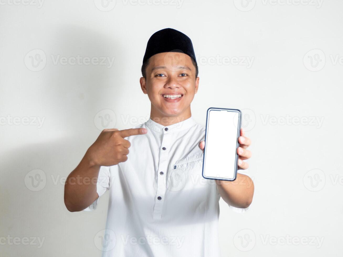 Asian muslim man smiling and showing mobile phone blank screen. Moslem wearing white shirt and black cap photo