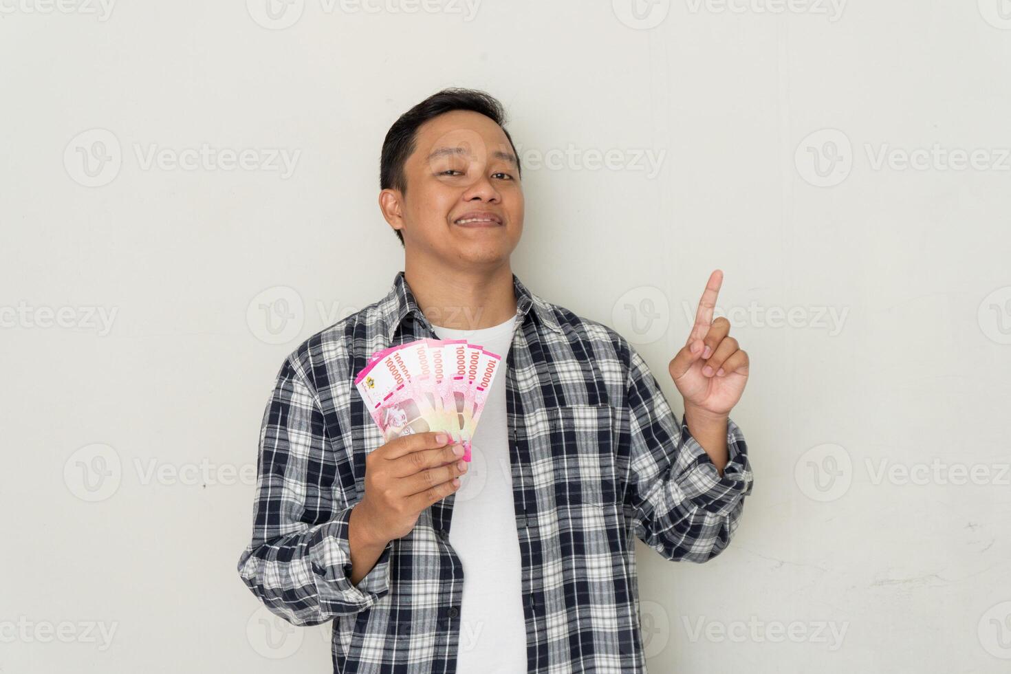 Asian guy smiling happy while holding mobile phone and bunch of money in Rupiah currency. photo
