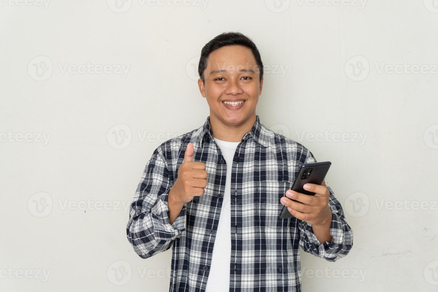 Young Asian man smiling happy and showing thumbs up gesture and smartphone in hand photo