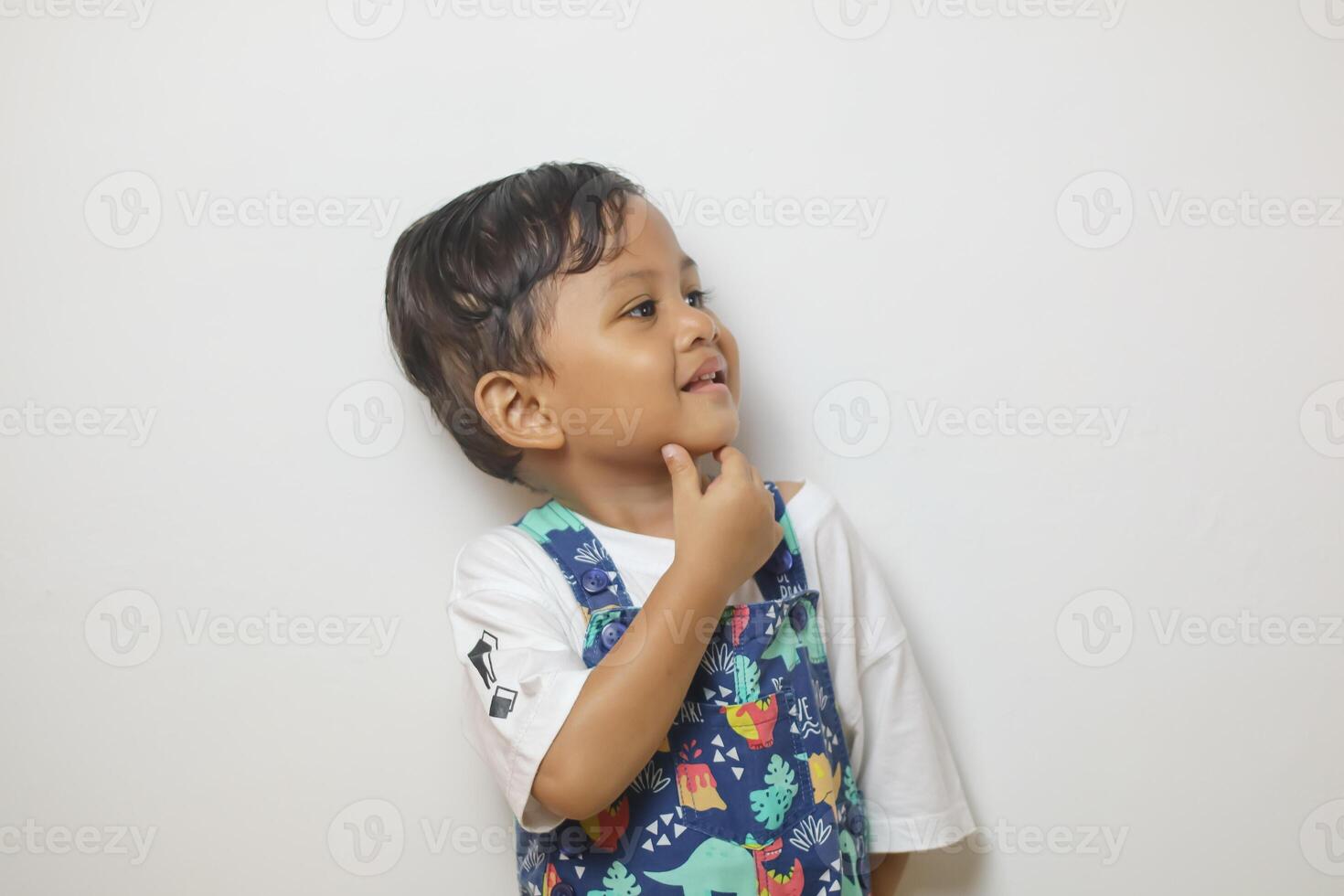 Thoughtful young boy with curly hair looking aside at copy space, touching chin in contemplation, wearing blue shirt on yellow background, embodying curiosity and deep thought photo
