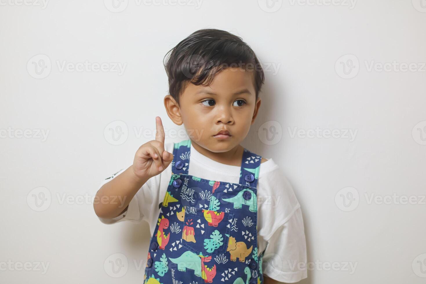 Cute smiling boy in plain white t shirt looking and pointing hand up in isolated studio light blue color background photo