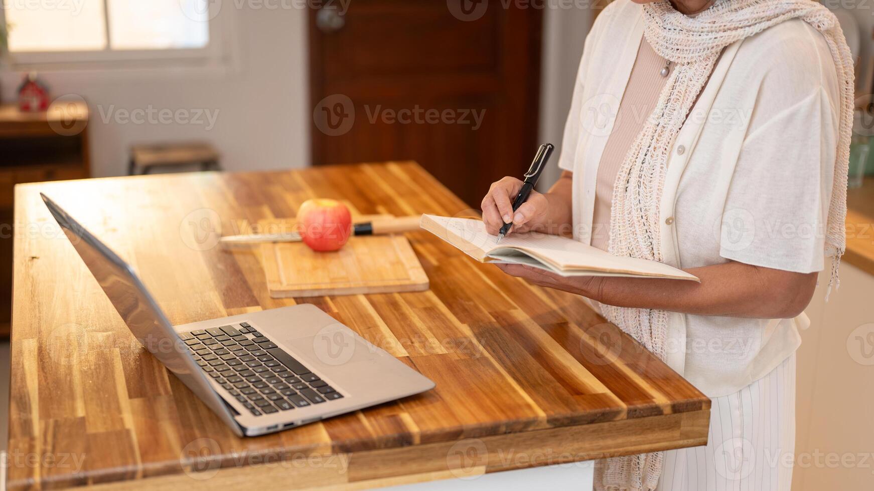 A retired woman writing down ingredients in a book while learning a new recipe on the internet. photo