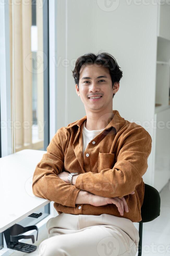 Handsome, positive Asian man sits at a table with his arms crossed in a room, smiling at the camera. photo