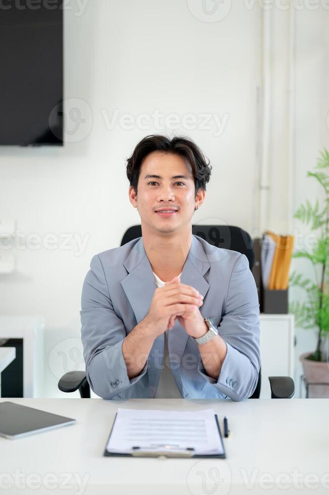 A successful Asian businessman sits at his desk in a modern office, smiling at the camera. photo