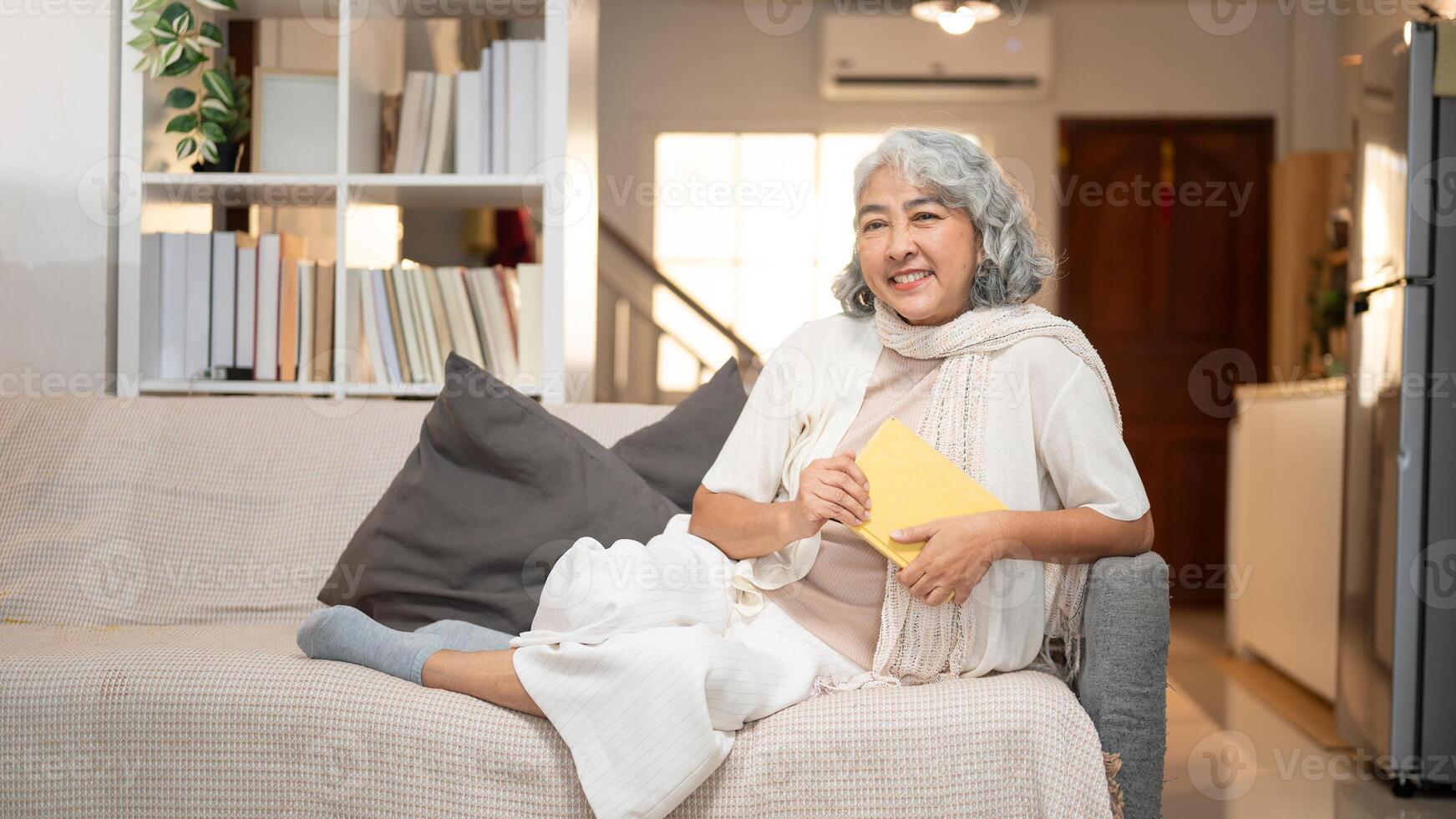 A happy Asian retired woman relaxes on a sofa, contemplating about happy life while reading a book. photo
