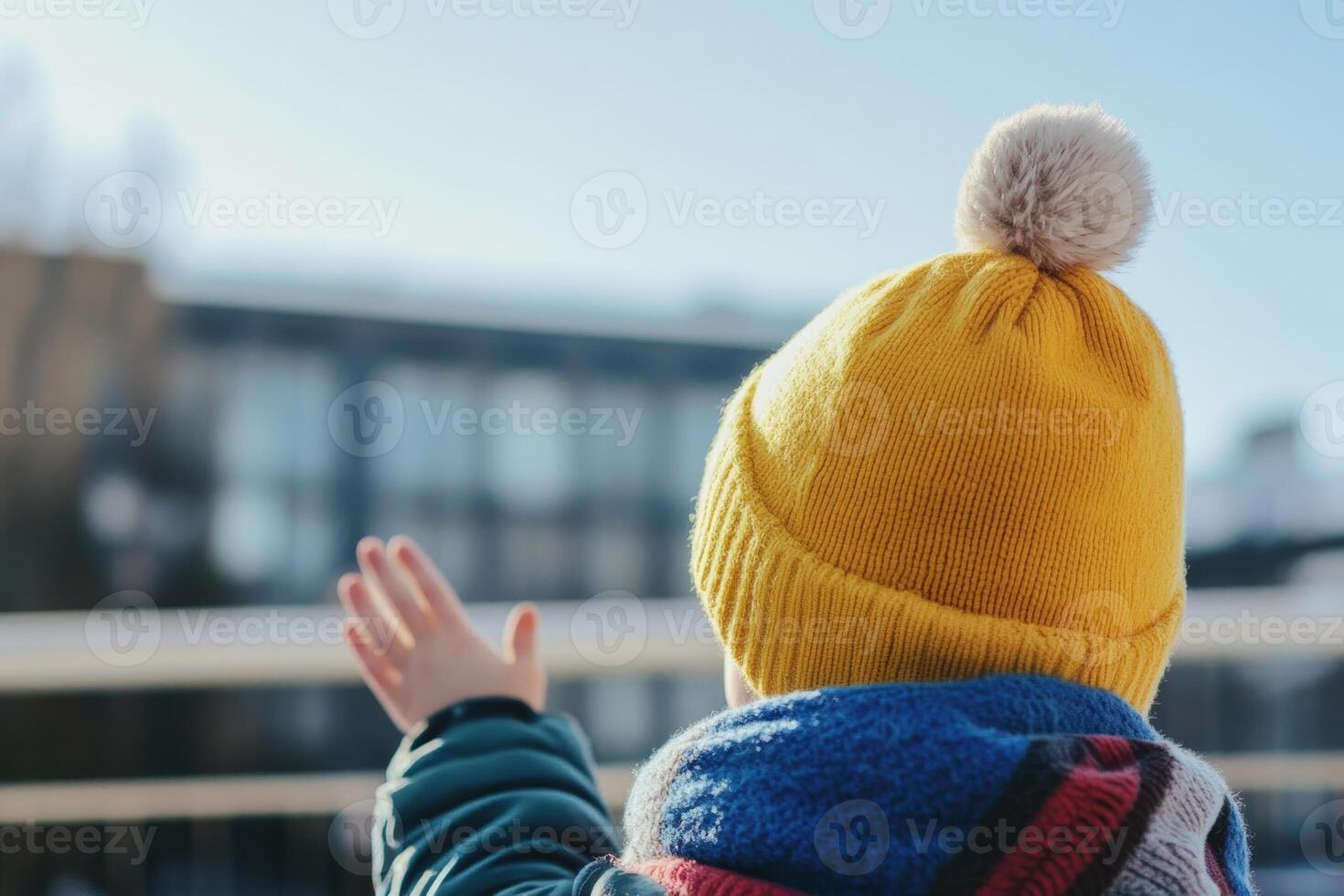 Child waves goodbye while wearing a bright yellow hat during a sunny winter day outside a building photo