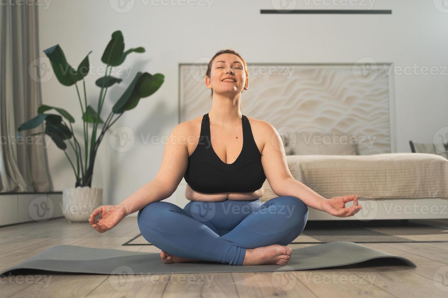 Yoga mindfulness meditation. Woman practicing yoga in living room at home. Plus size female sitting in lotus pose meditating smiling relaxing indoor. Girl doing breathing practice. Yoga at home photo