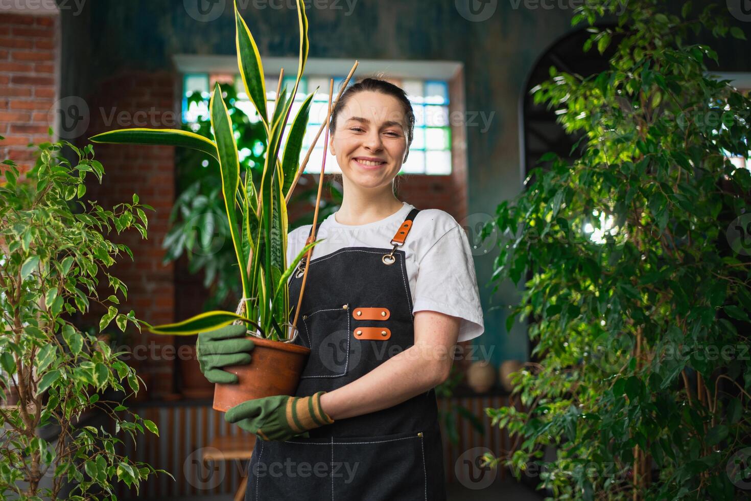 Running of own business. Young woman florist holding plant in pot wearing apron in botanical store. Happy small business owner working at flower shop smiling surrounded by plants. Small business photo