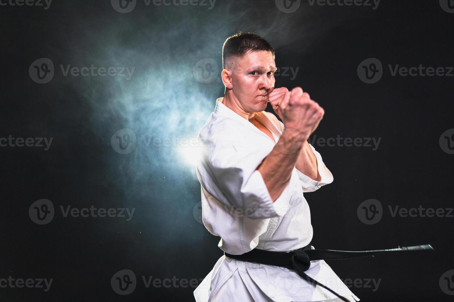 Strong man fighter in white kimono for karate martial arts with black belt preparing for fight training in black background. Man training moves punches kicks practicing shadow fight ready for sparring photo