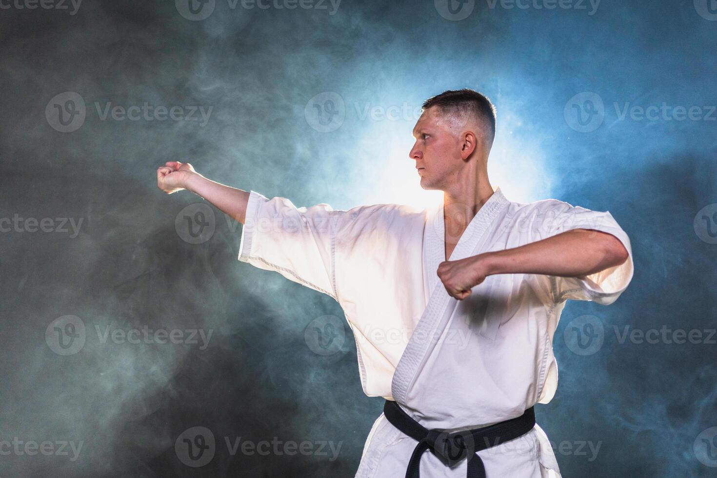 Strong man fighter in white kimono for karate martial arts with black belt preparing for fight training in black background. Man training moves punches kicks practicing shadow fight ready for sparring photo
