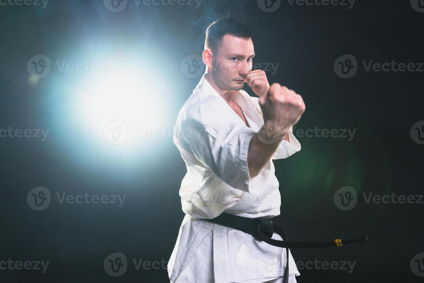 Strong man fighter in white kimono for karate martial arts with black belt preparing for fight training in black background. Man training moves punches kicks practicing shadow fight ready for sparring photo