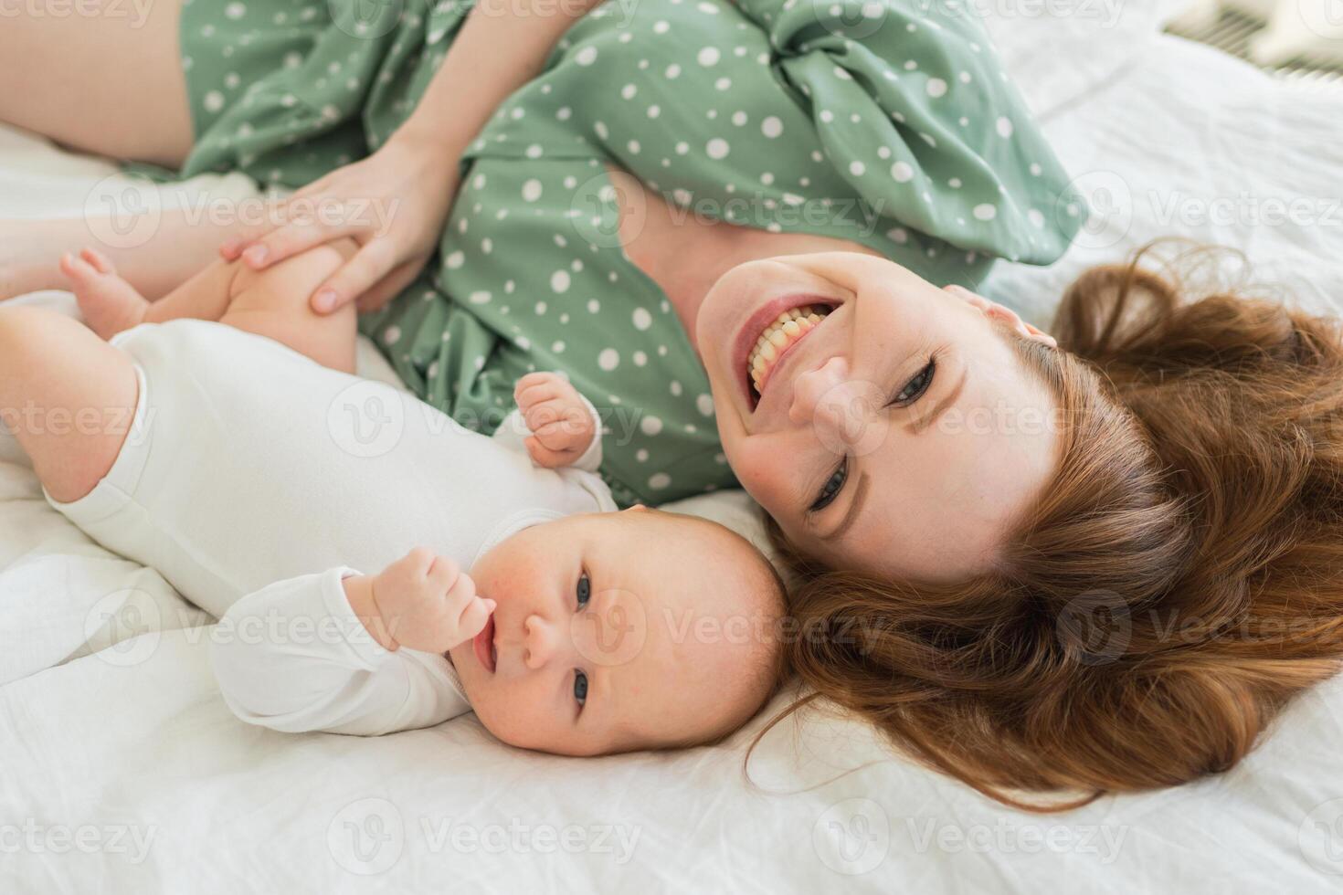 Happy family at home. Mother playing with little toddler child on bed. Mom baby relax playing having fun together. Mother looks at camera with love care. Mom of breast feeding baby rest in living room photo