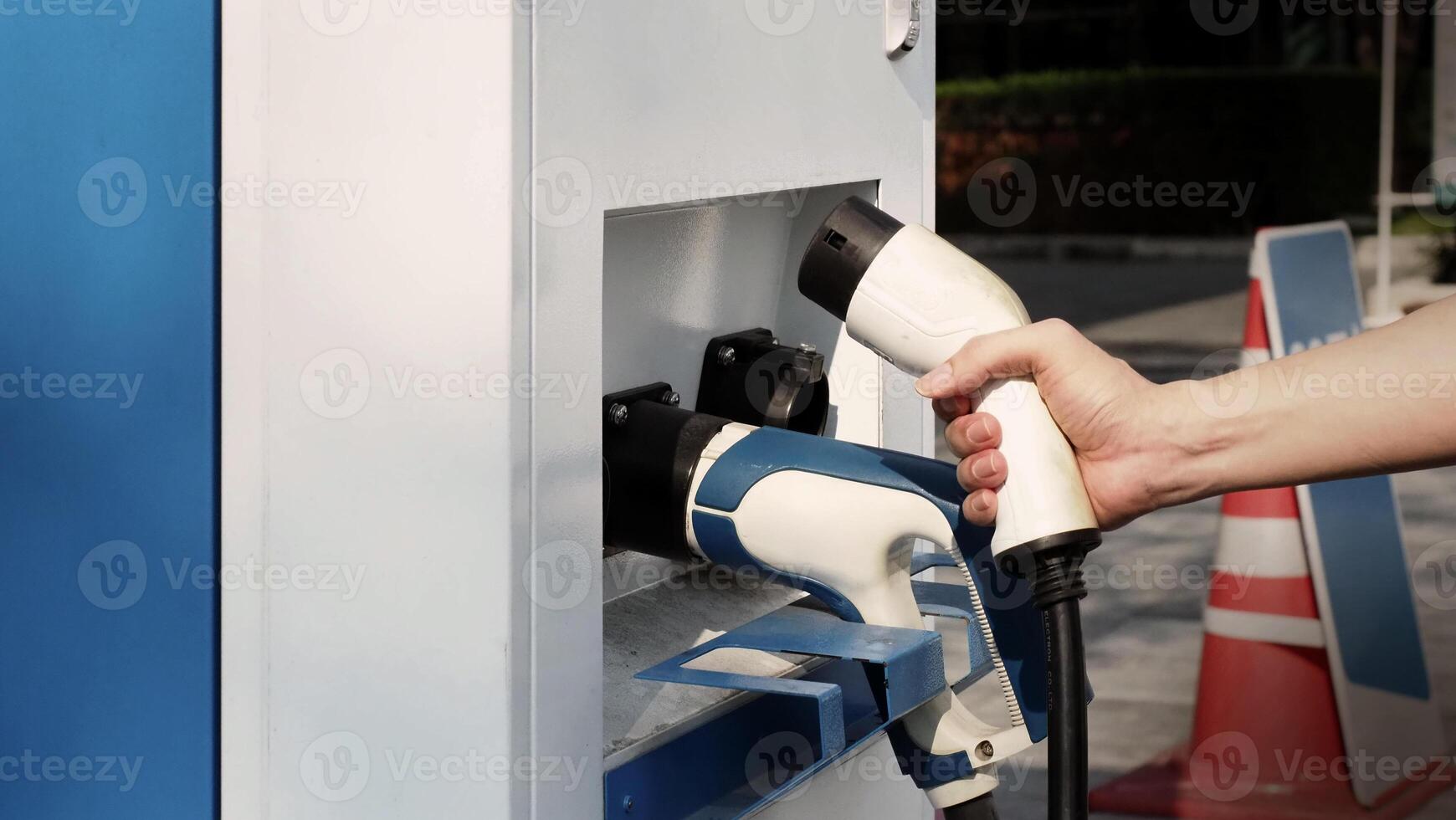 Driver of electric car hold batterie charger plug. Hand holding EV charging plug in front of camera with blurred background of outdoor. Sustainable energy powered electric charging station background photo