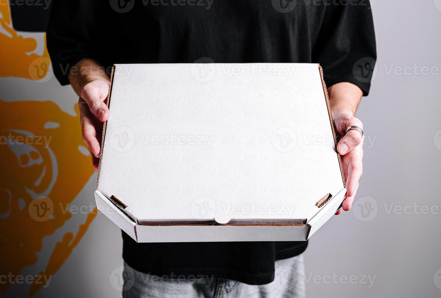Individual holding a blank white pizza box in a casual setting with a colorful wall background photo