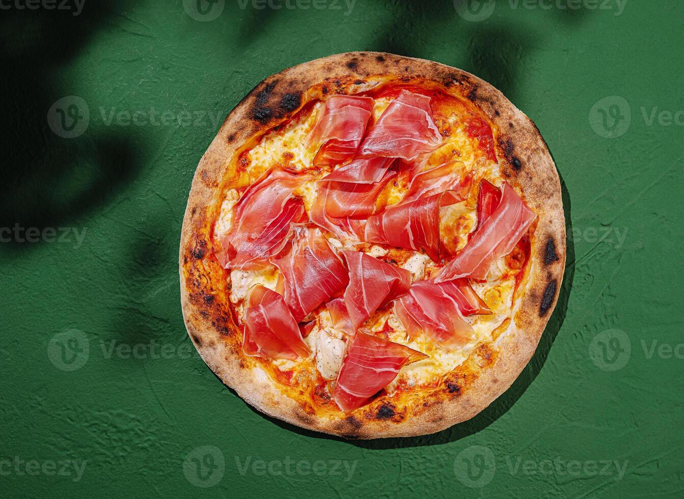 Freshly made pizza with prosciutto atop a rustic green background in bright lighting photo