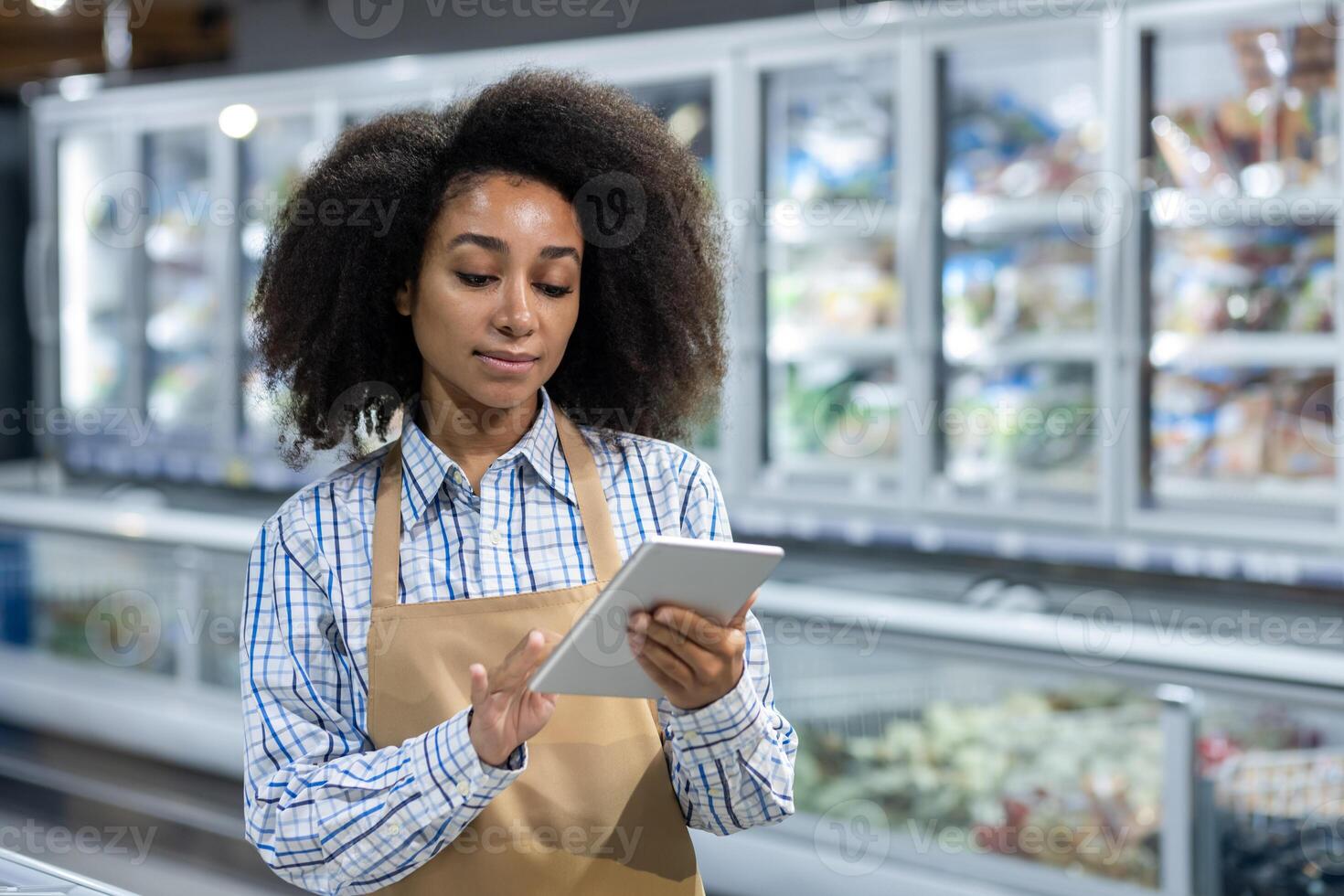 Supermarket employee wearing apron using digital tablet in frozen foods section. Professional woman managing inventory, order processing, and customer service. Concept of modern grocery store technology and efficiency. photo