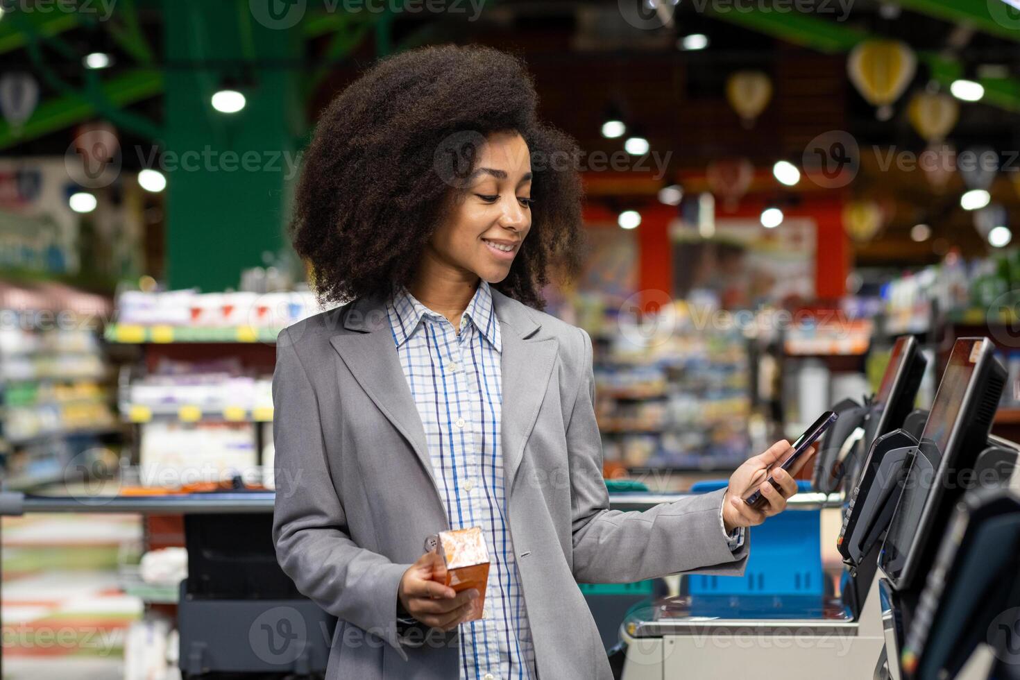 Smiling woman using smartphone at self-checkout kiosk in grocery store. Professional attire, holding beverage cup. Modern shopping experience, convenience, technology photo