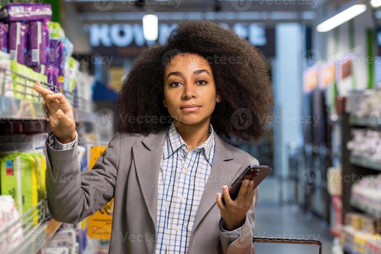 Confident woman in a supermarket aisle holding a smartphone and reaching for an item on the shelf. photo