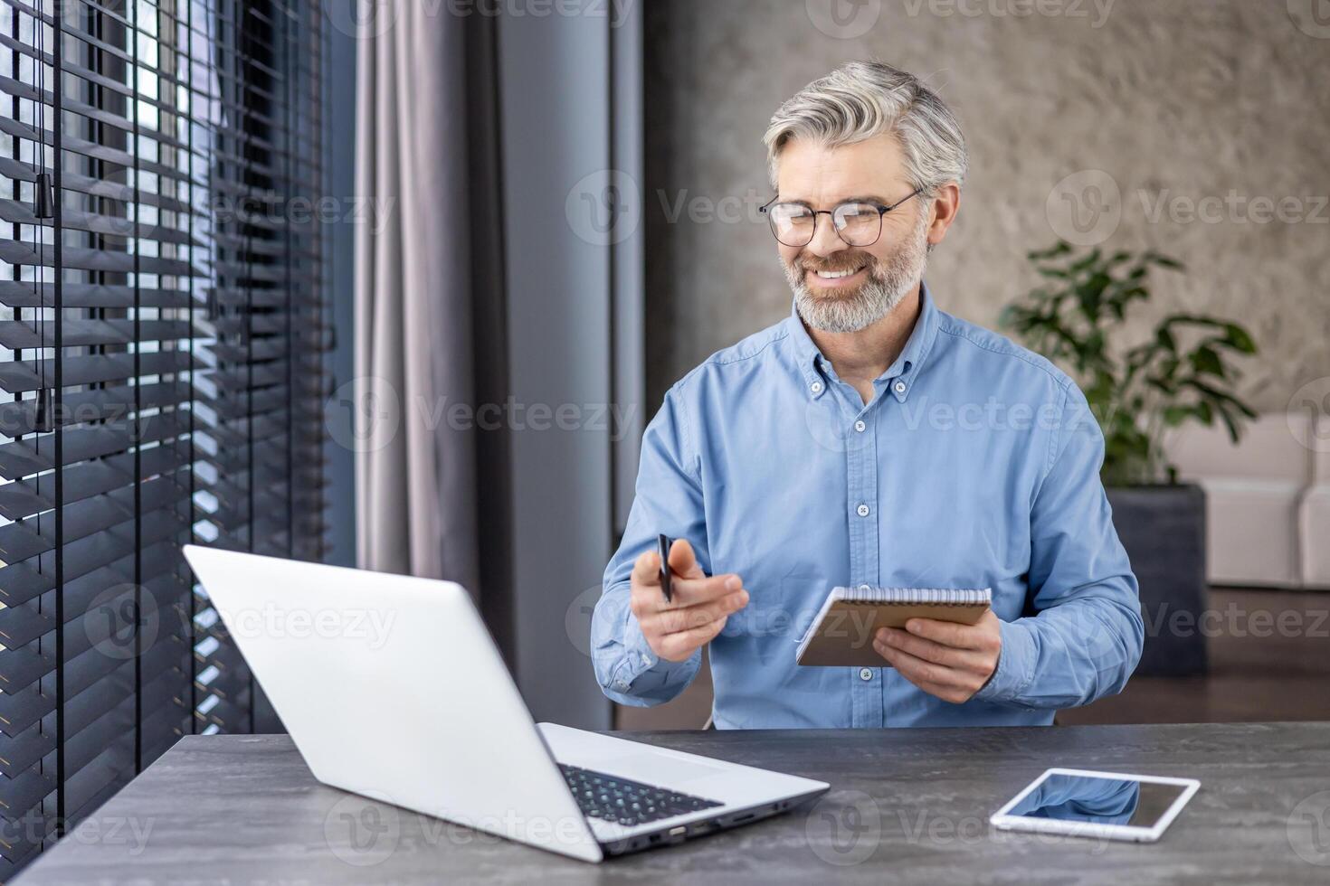 Senior tired man sits in the office in front of a laptop, talks, studies and works remotely online, holds and writes in a notebook. photo