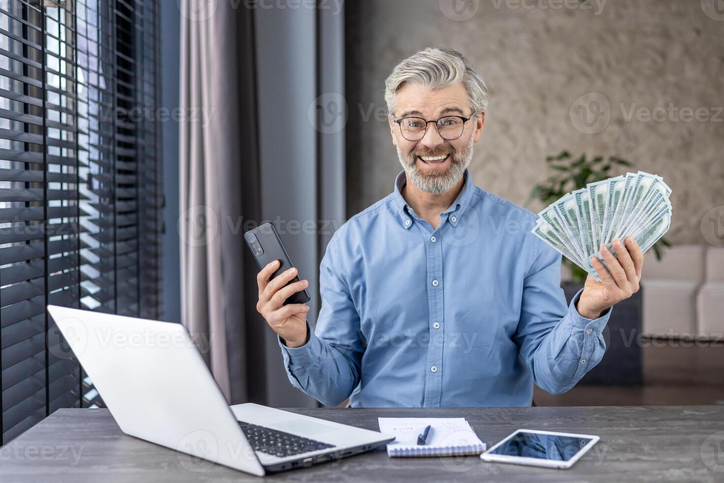 Businessman holding fan of dollar bills and smartphone while sitting in domestic office with portable computer. Joyful mature man getting excited about massive income after productive working day. photo
