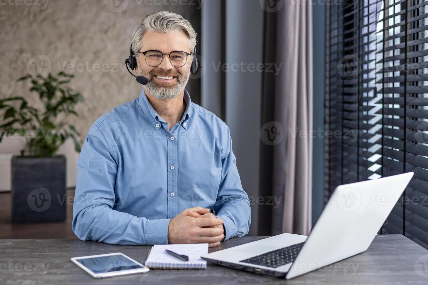 Portrait of mature successful gray haired man inside office at workplace inside office, businessman with headset phone and laptop for call smiling and looking at camera. photo