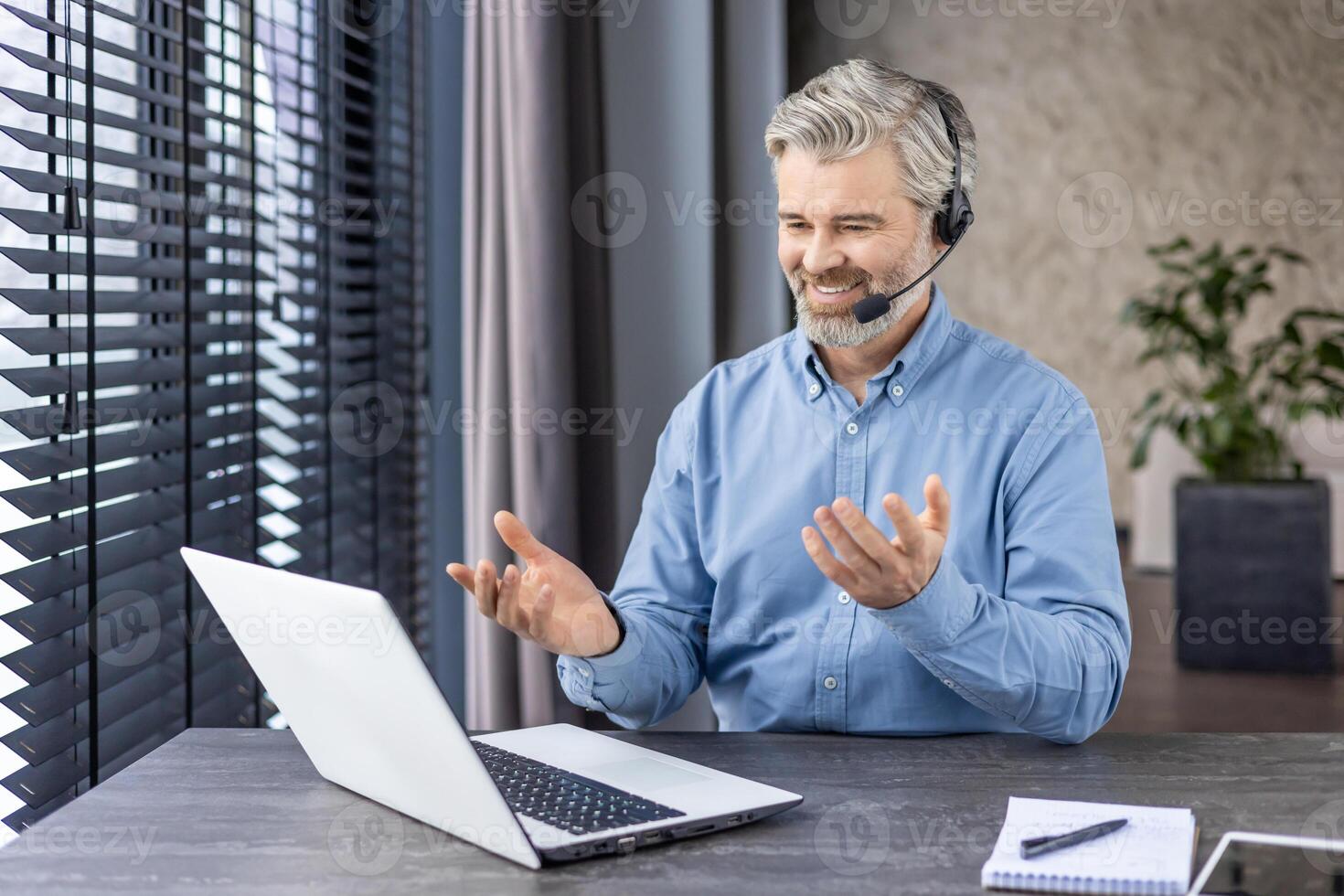 Elderly grey-haired man in denim shirt sitting at domestic workspace with laptop and having distant meeting with coworkers. Caucasian businessman in headset using online communication privileges. photo