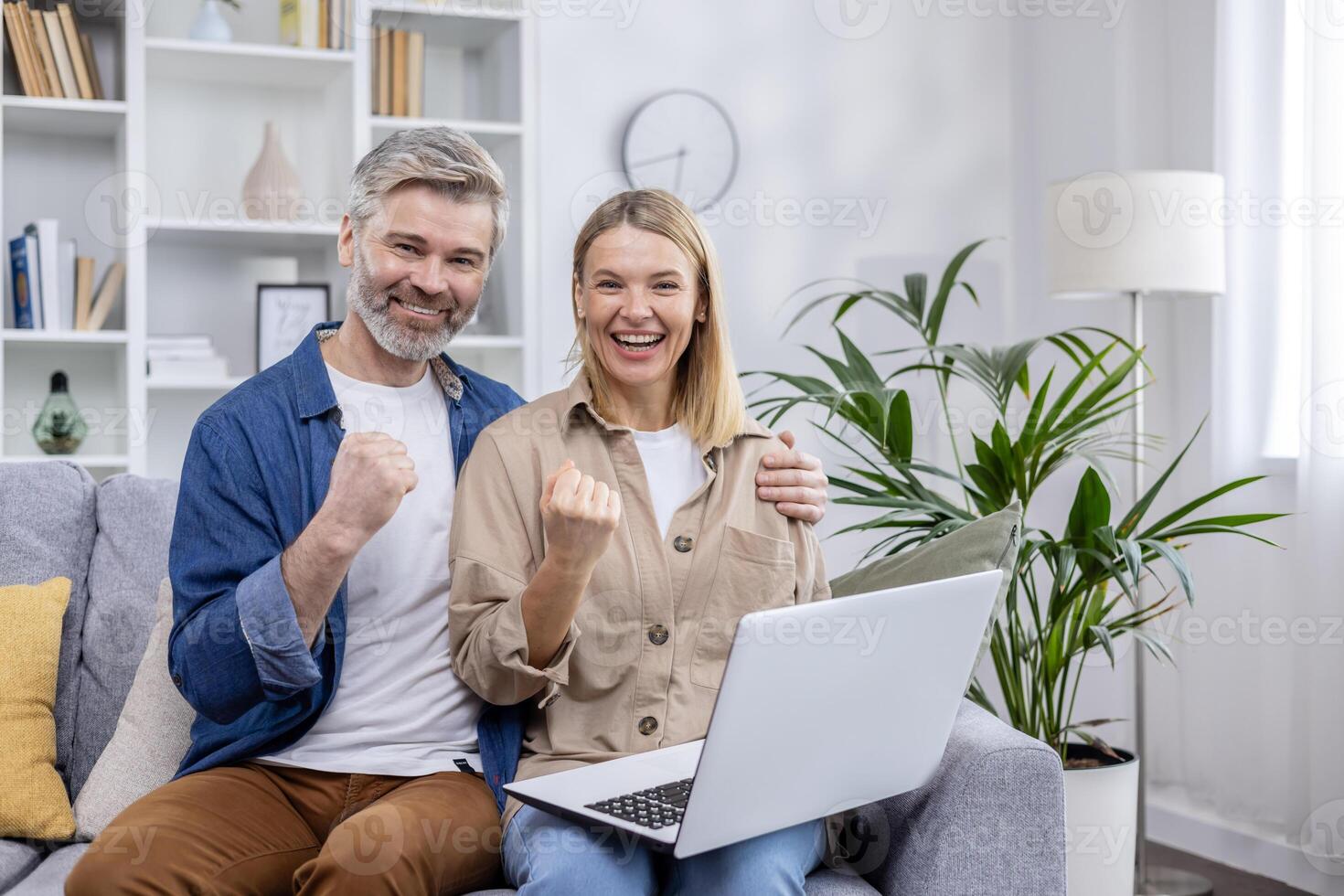 Joyful couple cheering while using a laptop, seated on sofa in cozy, modern living room. Expressing success and happiness. photo