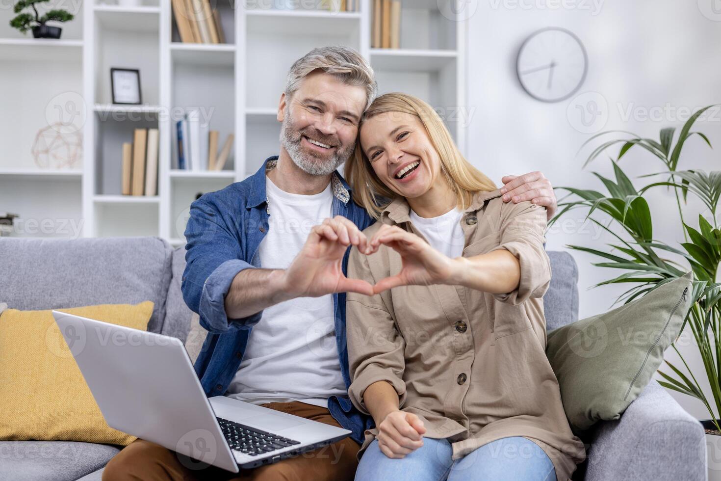 Cheerful mature couple making a heart shape with their hands while sitting on the couch at home. A perfect expression of love and partnership. photo