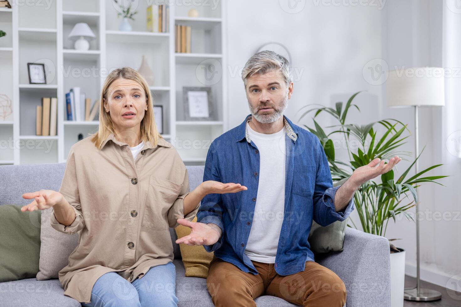 Middle-aged couple sitting on the sofa with confused expressions in a modern living room, conveying feelings of uncertainty and misunderstanding photo