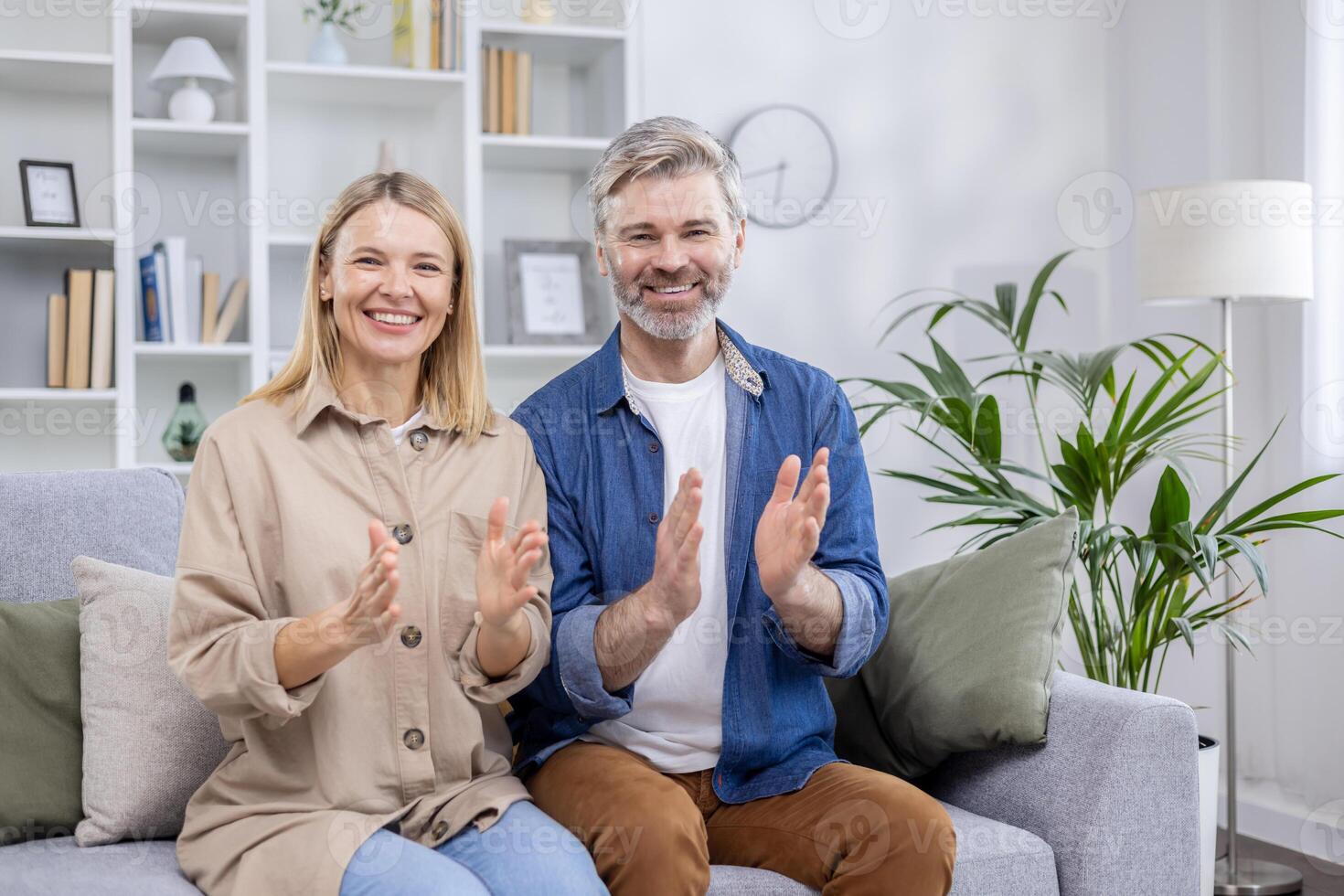 A joyful middle-aged couple sitting on a couch, clapping hands with broad smiles in a stylishly decorated living room. Expressing happiness and contentment. photo