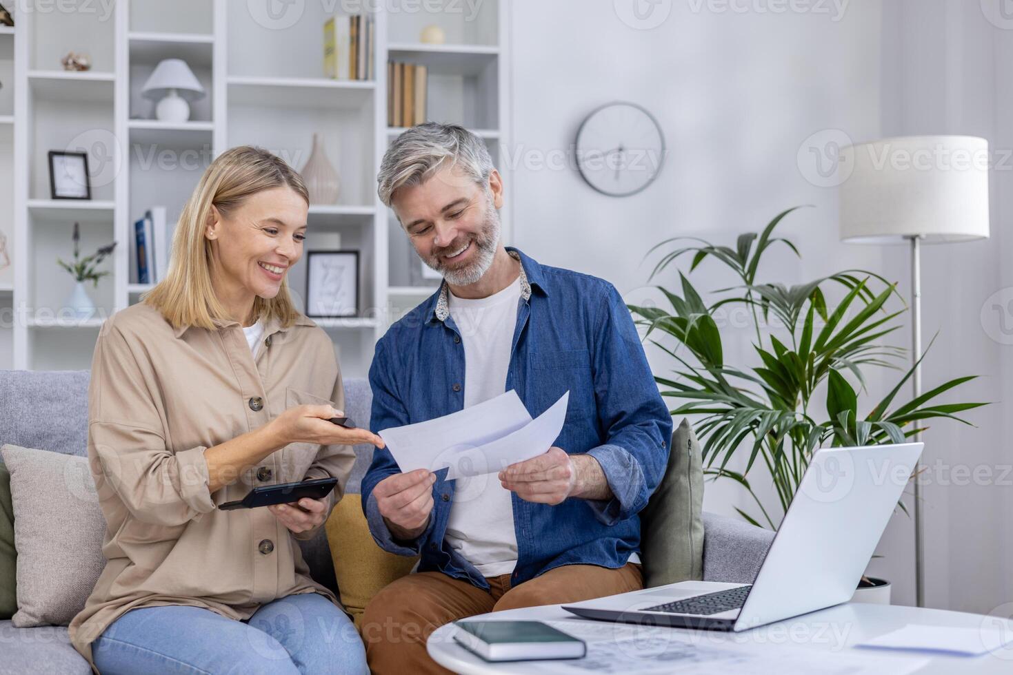 A mature couple in a cozy living room sorts and discusses documents with a laptop and calculator. They show teamwork and contentment while managing their finances. photo