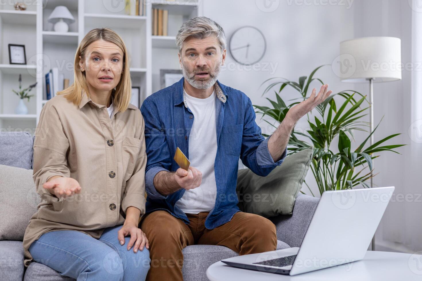 Middle-aged couple sitting on couch looking confused, holding a credit card, with laptop in front of them. Discussing uncertain financial issue or online payment problem. photo