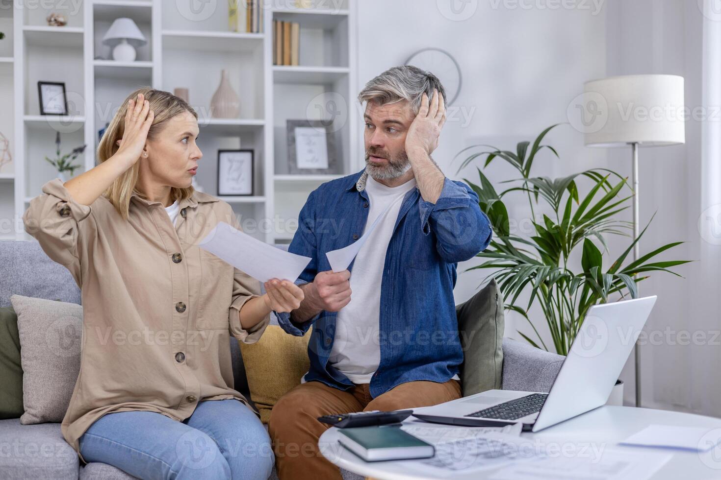 Stressed couple sitting on the couch discussing bills and finances. They appear worried while holding papers in their hands and looking at each other. photo