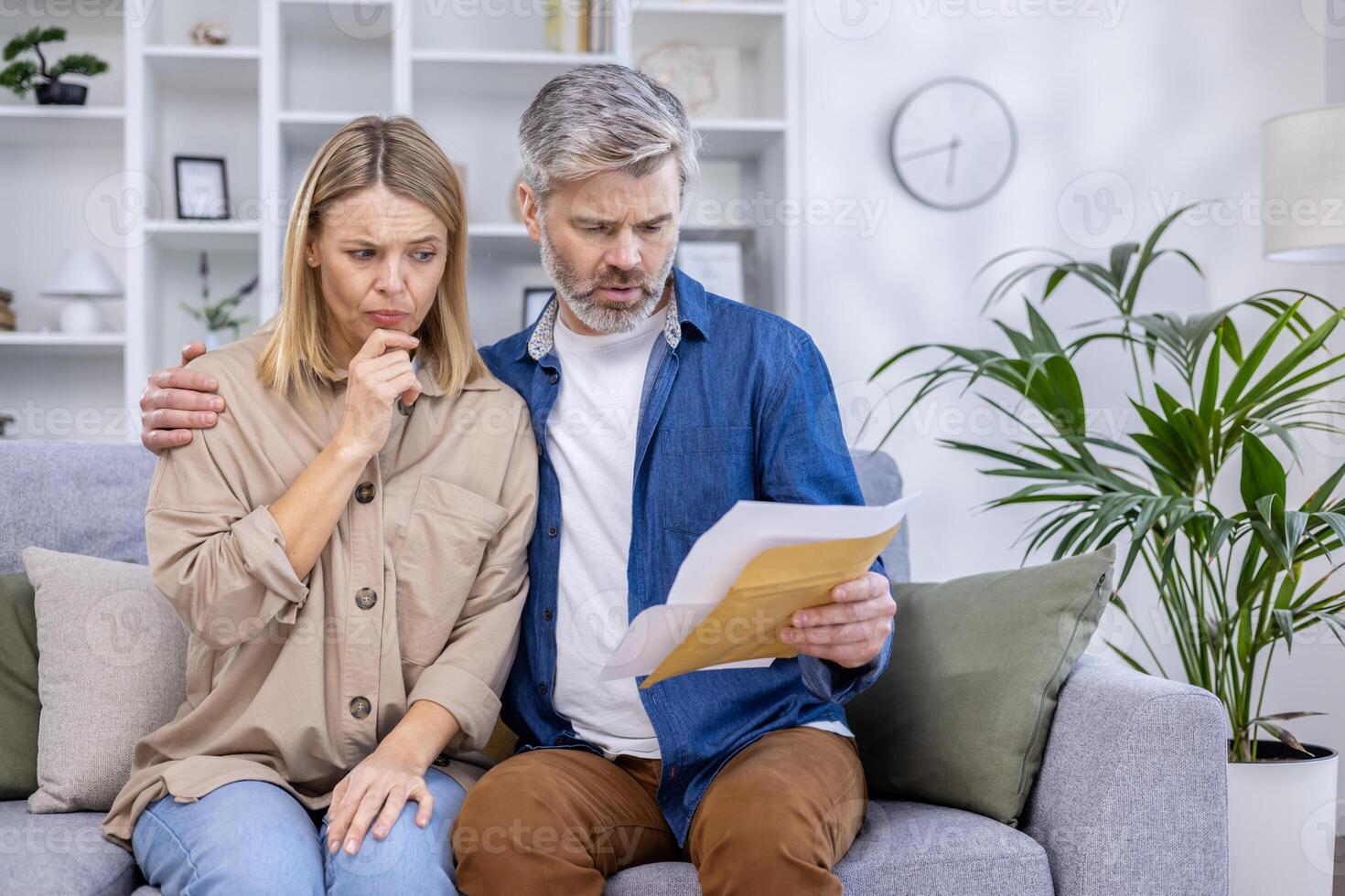 A concerned couple sitting on the couch, examining financial paperwork in their living room. They appear stressed while trying to understand the contents of the envelope. photo