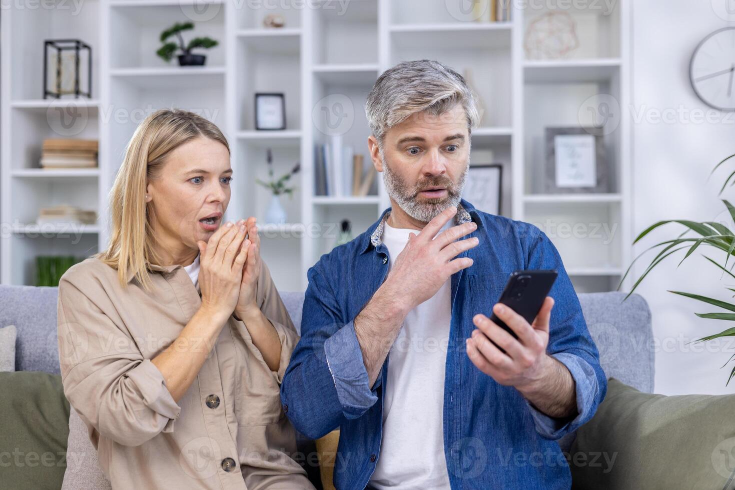 Middle-aged couple with shocked expressions, looking at a smartphone while sitting on the couch in a modern living room. photo