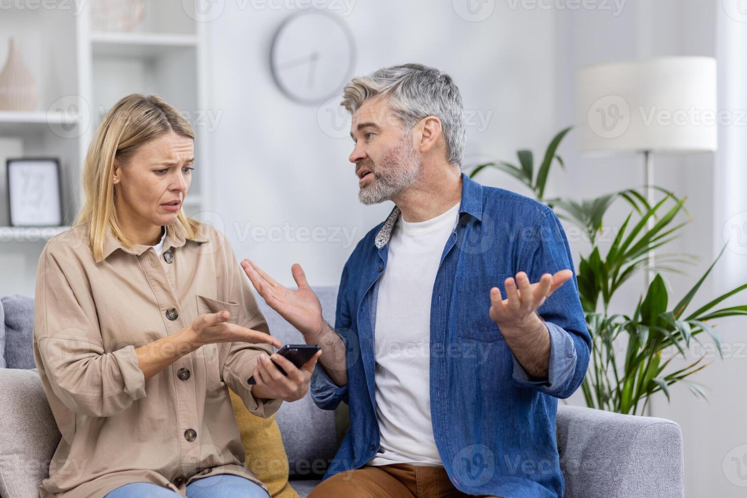 A couple seated on a couch engaged in an argument over a smartphone, depicting emotions of jealousy and conflict possibly due to social media. photo