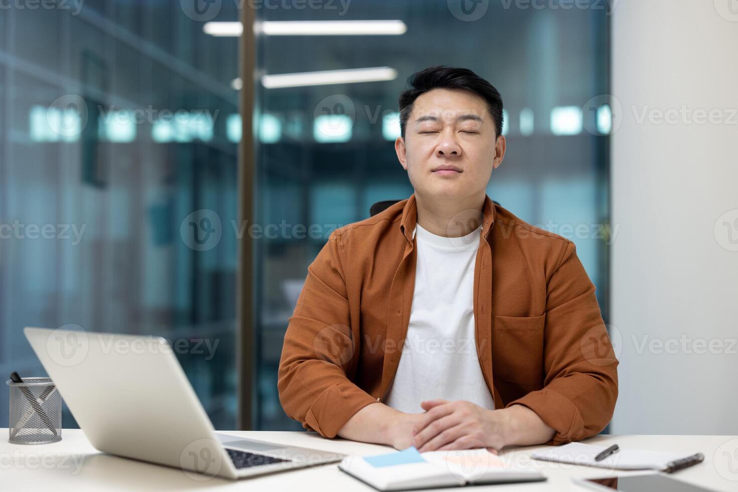 Asian man meditating at his desk in an office, practicing relaxation and mindfulness techniques amidst work. photo