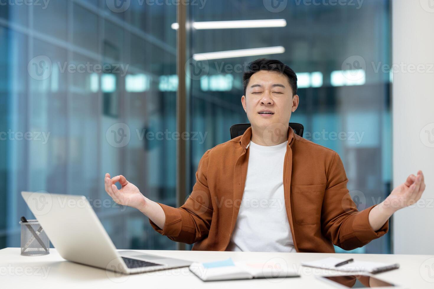 Mature Asian man resting meditating sitting in lotus position inside office. Businessman working with laptop at workplace, doing breathing exercises. photo
